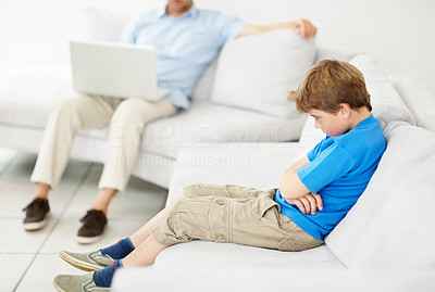 Sad little boy sitting on sofa with his father working on laptop in background
