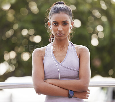 Buy stock photo Portrait of a sporty young woman wearing earphones and standing with her arms crossed while exercising outdoors