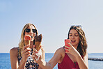 Two friends taking photos of ice cream eating on beach using smart phone technology on adventure travel