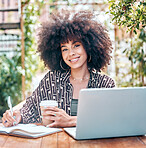 Young happy mixed race businesswoman with a curly afro writing in a notebook drinking coffee and working on a laptop sitting outside at a cafe. Hispanic female student studying at a restaurant

