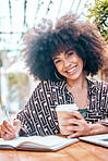 Smiling african american freelancer with curly afro working remotely in a cafe during the day. Mixed race entrepreneur writing notes in a book while enjoying a takeaway coffee. Hispanic businesswoman