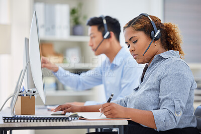 Buy stock photo Focused young mixed race female call centre telemarketing agent writing notes and talking on a headset while working on a computer in an office. Serious african american businesswoman consultant operating helpdesk for customer service support