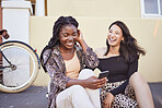 Best friends texting on smartphone while sitting on street.Two multiethnic female friends sitting on sidewalk and laughing while browsing social media.