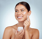 Smiling beautiful woman applying face cream while posing with copyspace. Caucasian model isolated against grey studio background with product on cheek. Moisturise and sunscreen for healthy skincare