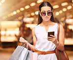 A young caucasian woman wearing sunglasses using a cell phone while carrying bags during a shopping spree. Young brunette woman sending a text with her smartphone after shopping in a mall. A little retail therapy is never a bad idea