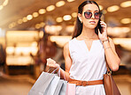 A young caucasian woman wearing sunglasses using a cell phone while carrying bags during a shopping spree. Young brunette woman on a call with her smartphone while shopping in a mall. A little retail therapy is never a bad idea