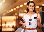 A young mixed race woman using a cell phone and bank card while carrying bags during a shopping spree. Young brunette woman purchasing items online with her smartphone and credit card in a shopping mall. A little retail therapy is never a bad idea
