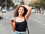 One happy beautiful young mixed race woman exploring the city while holding a takeaway coffee and touching her curly brunette hair. Hispanic travelling woman enjoying the view downtown on the weekend