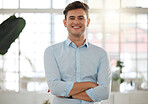 Young caucasian businessman standing with his arms crossed in an office alone at work. One happy male businessperson smiling while standing at work