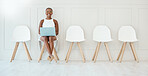 Portrait of a young focused African american businesswoman working on laptop while waiting for interview sitting on a chair against a white wall. Waiting for an interview in an office