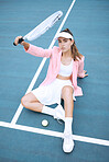 Trendy young tennis player wearing a pink jacket while sitting on a tennis court. Young hispanic sportswoman sitting with a tennis racket and ball on a sunny day