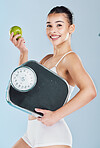Portrait of a young mixed race fit woman holding an apple and scale while posing against blue copyspace background. One athletic female showing the benefits of a healthy lifestyle and balanced diet
