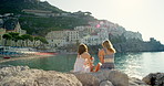 Happy friends taking photos with a cellphone while sitting by the seaside. Two women on holiday taking photos with a smartphone
